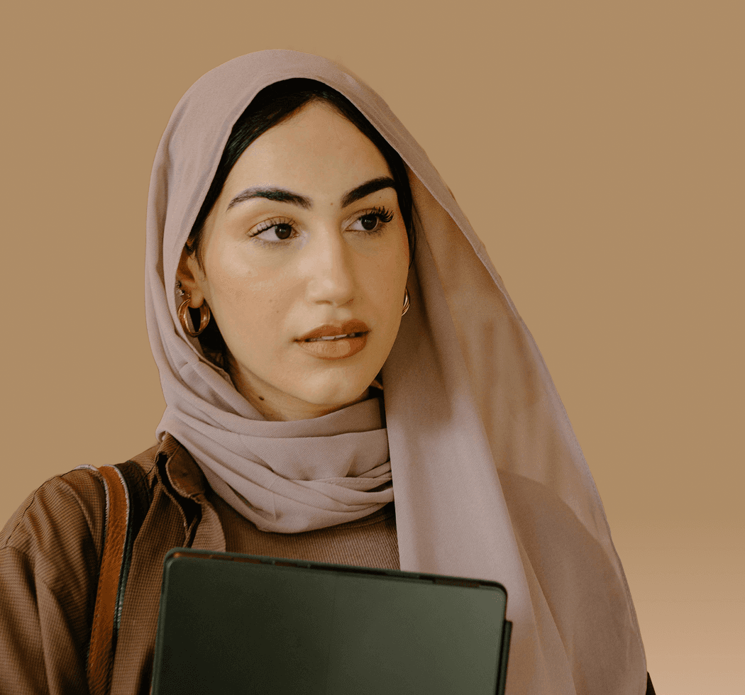 A person wearing a light pink hijab and brown top holding a tablet against a neutral beige background.
