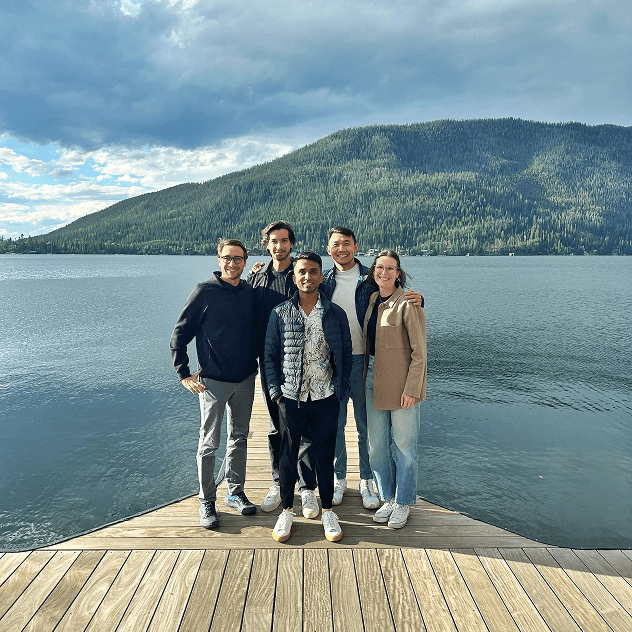 A group of five people standing on a wooden dock overlooking a serene lake with forested mountains in the background.