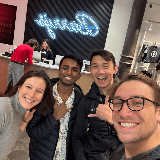 A selfie of five smiling people inside what appears to be a retail store with a blue neon \'Beauty\' sign visible in the background.